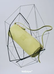 Light green cylindrical shoulder bag resting inside a geometric black wireframe sculpture against a grey background.
