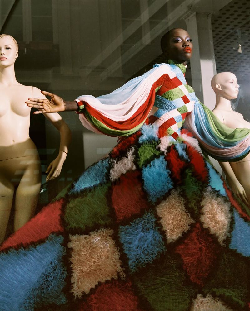 African-American woman in colorful dress striking a pose near mannequin in window display, fashion photography, dramatic lighting, editorial.