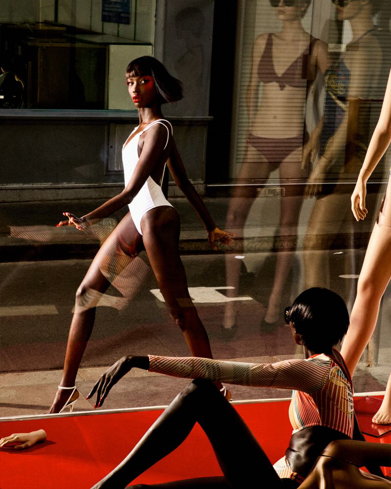 Dark-skinned woman in a white one-piece bathing suit strikes a pose in front of a store window with mannequin reflections.