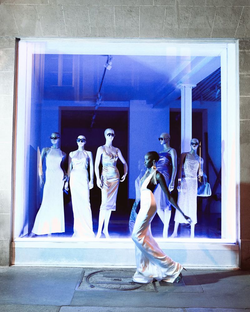 Woman in white gown walking by window display of mannequins in evening dresses with sunglasses, city street and building facade.