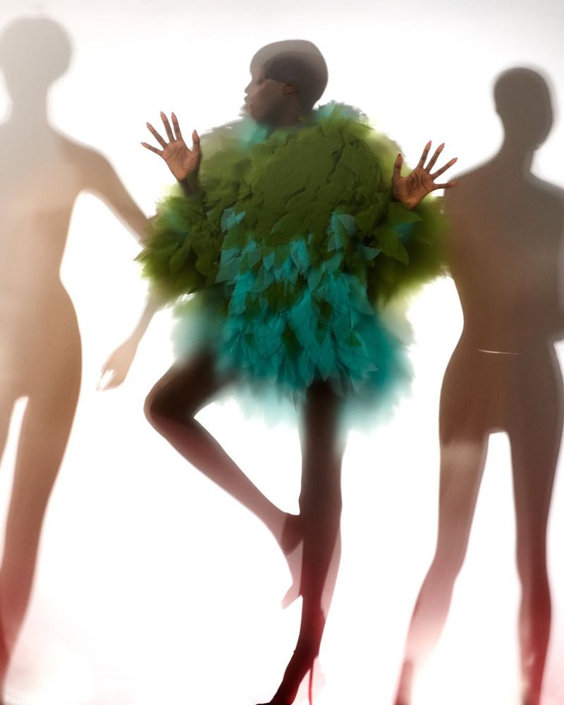 An adult female model with dark skin and a shaved head is posing in a bright studio, wearing a green and blue feathery coat.
