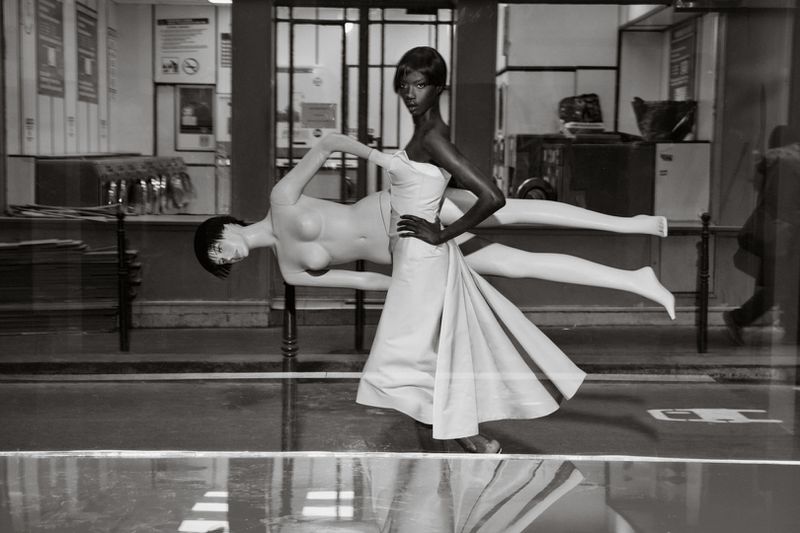 Black and white fashion shot of a Black woman in a dress standing next to a mannequin in a shop window, captured in medium shot.