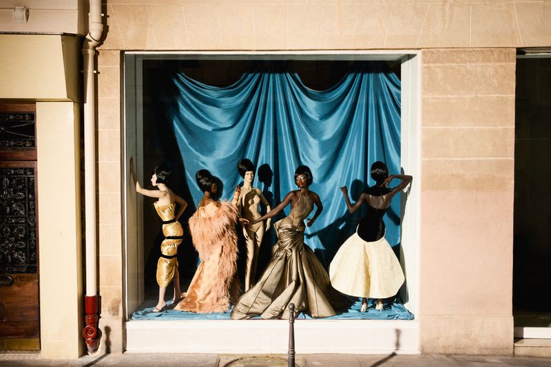 Fashion mannequins in gowns displayed in a storefront window with a blue draped background, showcasing an editorial fashion concept.