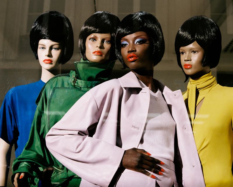 Fashion shot of a Black woman posing with three mannequin heads with black wigs wearing colorful tops and jackets in a studio.
