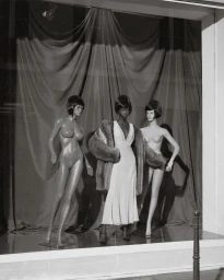 Three mannequins inside a shop window display, featuring a central dark-skinned mannequin in a white dress and fur coat