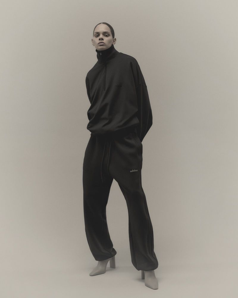 A person in a black tracksuit with grey boots stands in a studio. The individual is centered in the frame against a neutral backdrop.