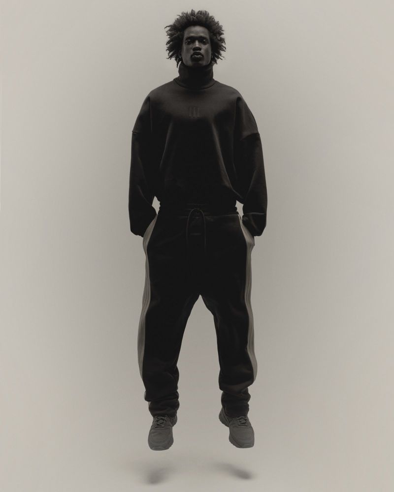 A male model with an afro poses mid-air in a black turtleneck and sweatpants with grey stripes against a neutral studio backdrop.