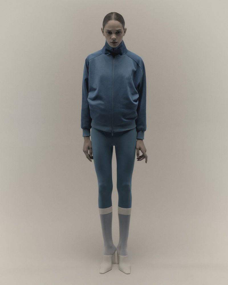Model in a blue athletic zip jacket and leggings standing against a neutral studio background