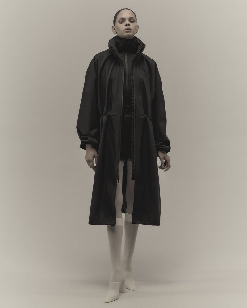 A slender fashion model poses in a studio wearing a long black zippered jacket over a dark turtleneck and opaque tights with white heeled shoes.
