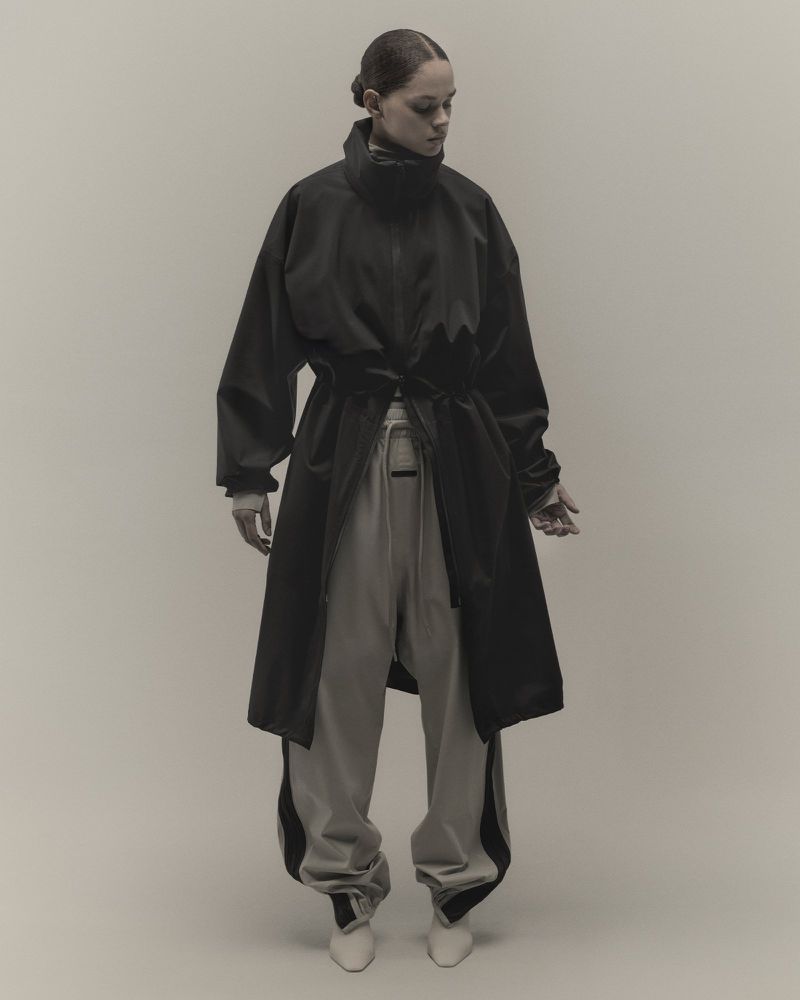 Woman in stylish black coat and grey pants posing against a neutral background in a fashion-forward, editorial-style shot.