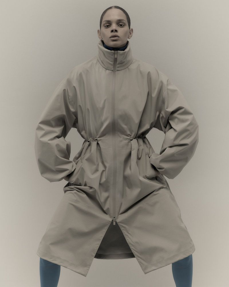 Fashion portrait of a woman in a beige trench coat with large sleeves against a plain backdrop in a studio environment.