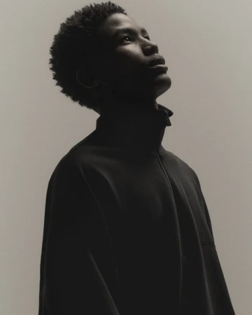 Portrait of an adult male wearing a black jacket looking upward, set against a light neutral background in an indoor studio environment.