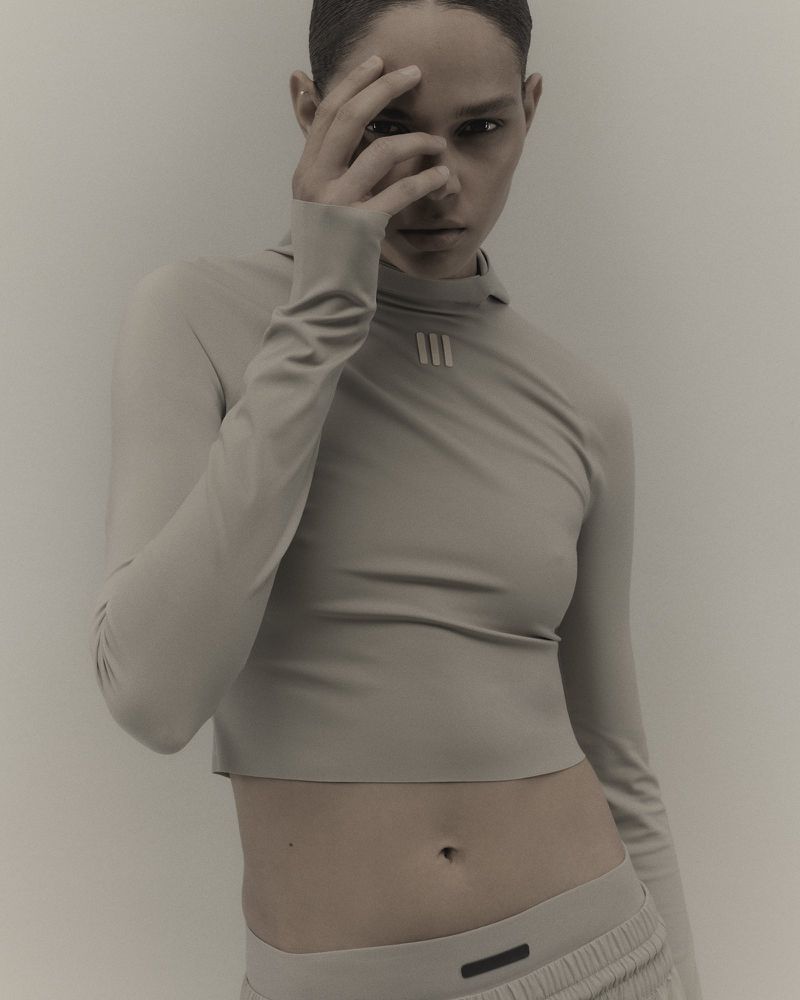 Adult female fashion model in long-sleeved crop top and sweatpants poses indoors with a hand near her face in a studio setting.