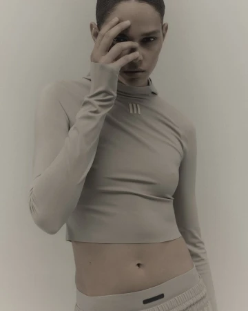 Adult female fashion model in long-sleeved crop top and sweatpants poses indoors with a hand near her face in a studio setting.