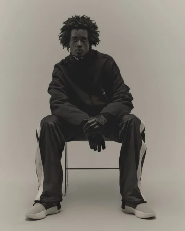 Portrait of a Black male fashion model sitting on a chair in a photo studio, wearing black tracksuit, leather gloves, and grey sneakers.