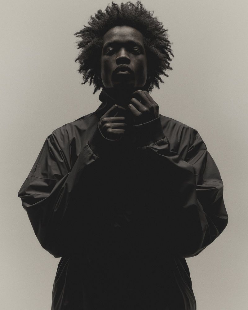 Portrait of an adult man with curly hair, wearing a dark jacket in a studio setting. The subject is looking directly at the camera.