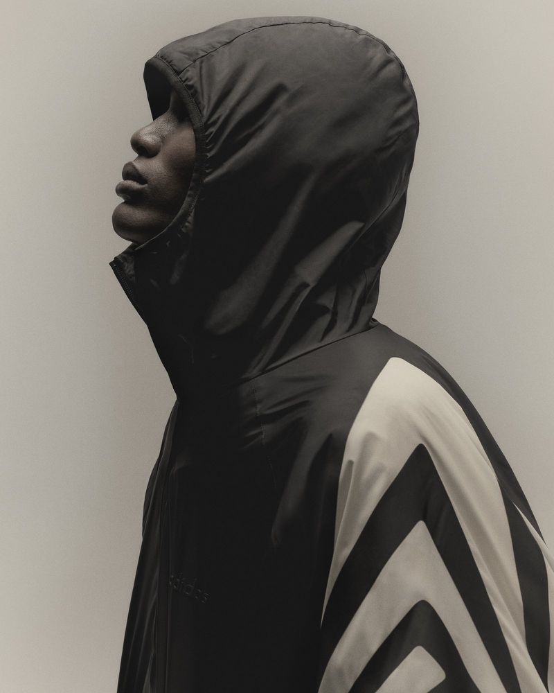 Side profile of an African American adult male wearing a black hooded jacket with white chevron sleeve details, set against a neutral backdrop.