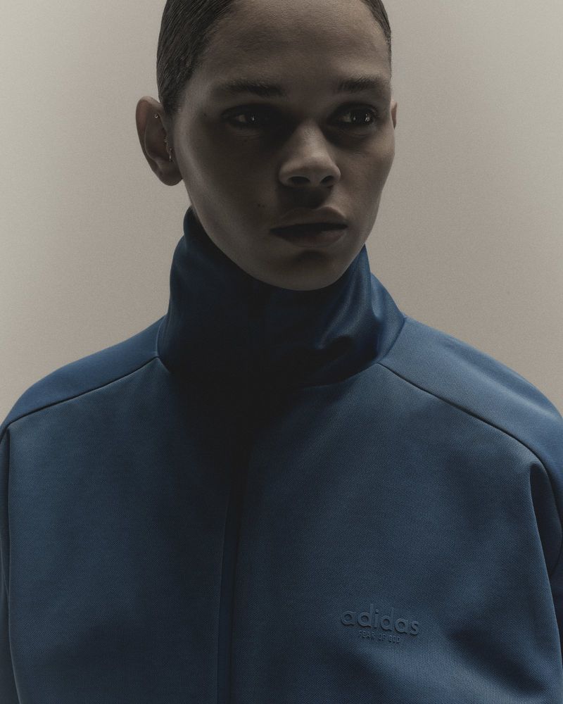Close-up portrait of a model wearing a high-collared blue athletic jacket against a neutral grey background.