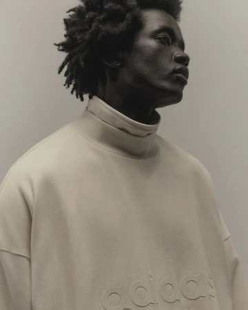 Portrait of an African-American man with dark curly hair wearing a beige high-neck sweatshirt, captured in a photo studio setting.