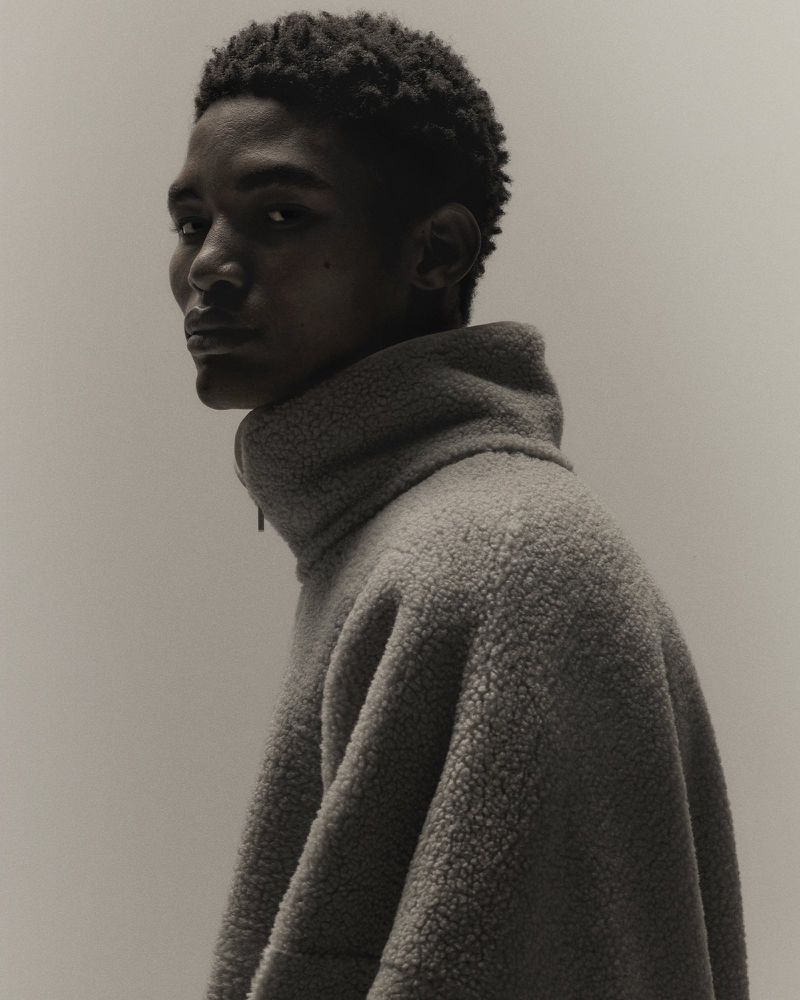 Portrait of a young adult male with dark hair, wearing a high-necked gray fleece sweater, looking over his shoulder against a neutral background.