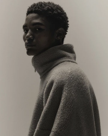 Portrait of a young adult male with dark hair, wearing a high-necked gray fleece sweater, looking over his shoulder against a neutral background.