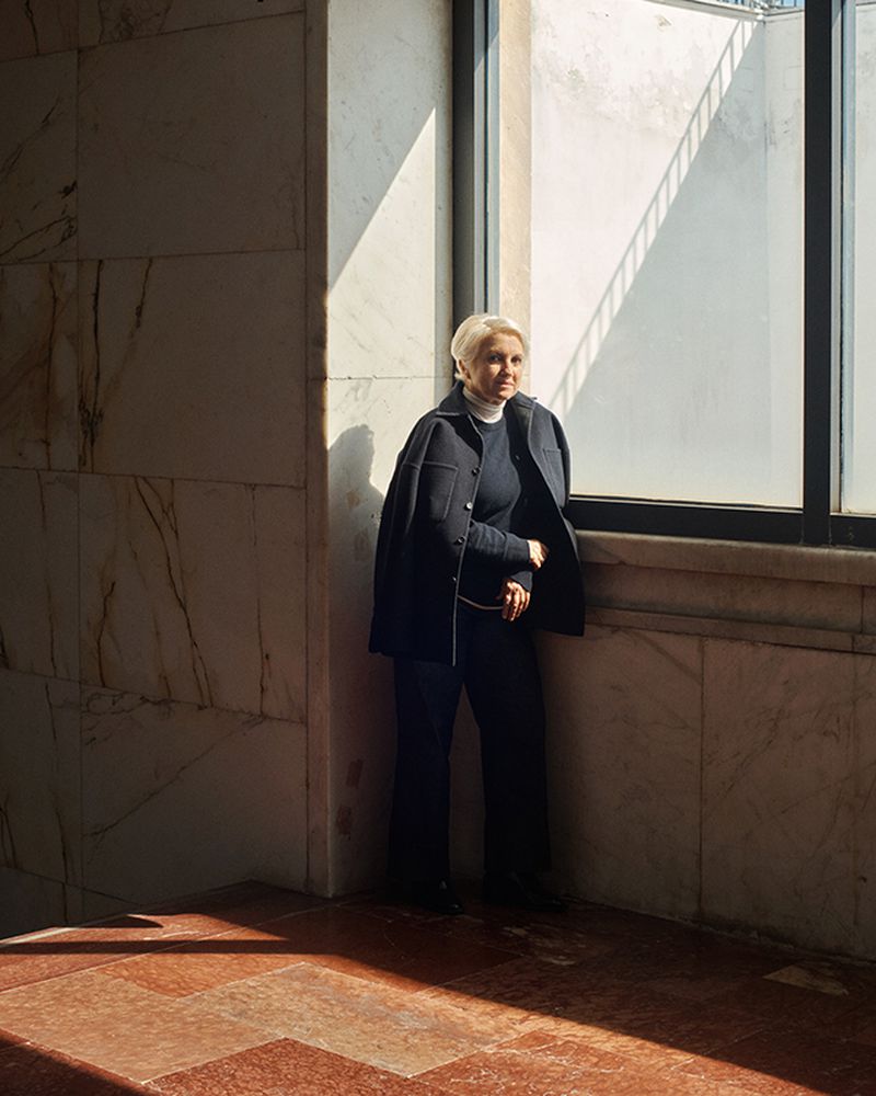 An older woman with short blonde hair stands near a window in a marble-walled building, wearing a navy jacket and pants, gazing outside.