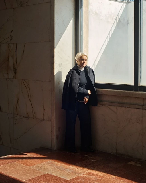 An older woman with short blonde hair stands near a window in a marble-walled building, wearing a navy jacket and pants, gazing outside.