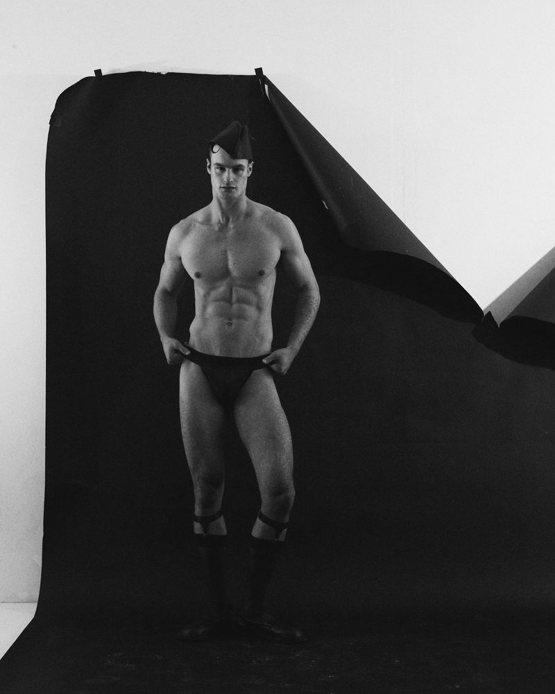 Monochrome portrait of a muscular adult male in posing in underwear and garrison cap, with a torn black paper backdrop.