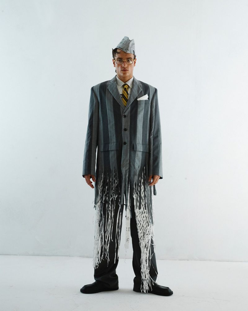 Man in an upcycled suit, made of paper and newspaper, accessorized with glasses, a tie, and black shoes, posing in a white studio.