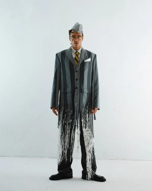 Man in an upcycled suit, made of paper and newspaper, accessorized with glasses, a tie, and black shoes, posing in a white studio.
