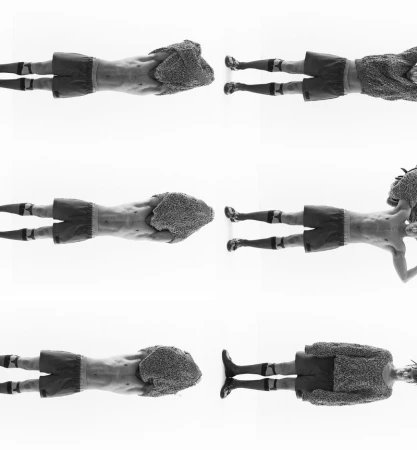 Monochrome studio shot: Six adult men lie horizontally, facing opposite directions, wearing shorts, socks, and boots. One wears a fur coat.
