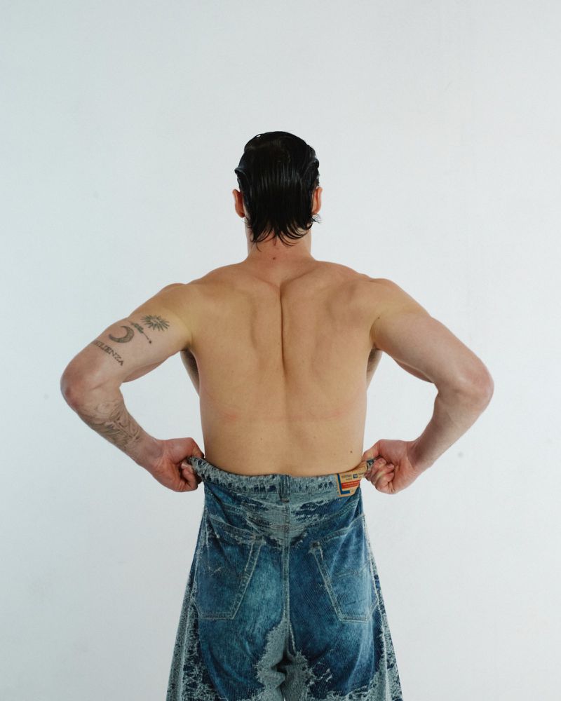 From behind, a muscular adult male wears blue denim jeans low, standing against a plain white backdrop in a studio setting.