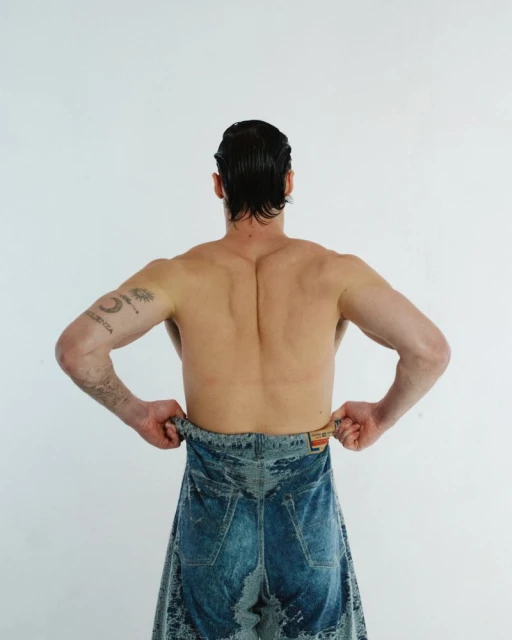 From behind, a muscular adult male wears blue denim jeans low, standing against a plain white backdrop in a studio setting.