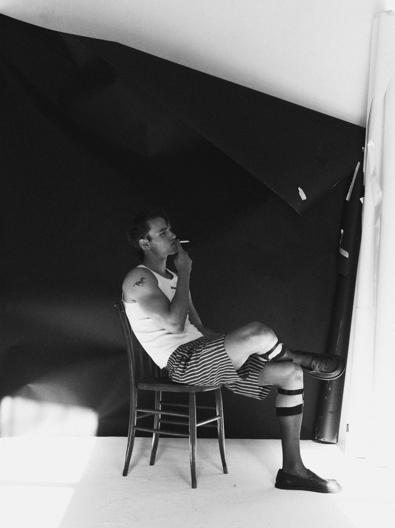 Black and white photo of a man sitting on a chair, smoking, with striped shorts, dark socks, and a dark background in a studio setting.