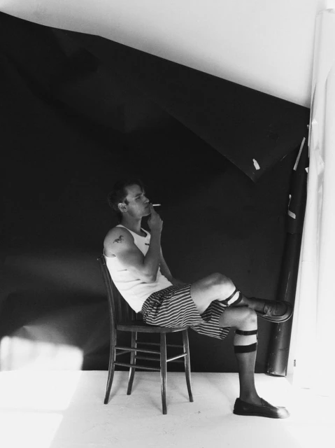 Black and white photo of a man sitting on a chair, smoking, with striped shorts, dark socks, and a dark background in a studio setting.