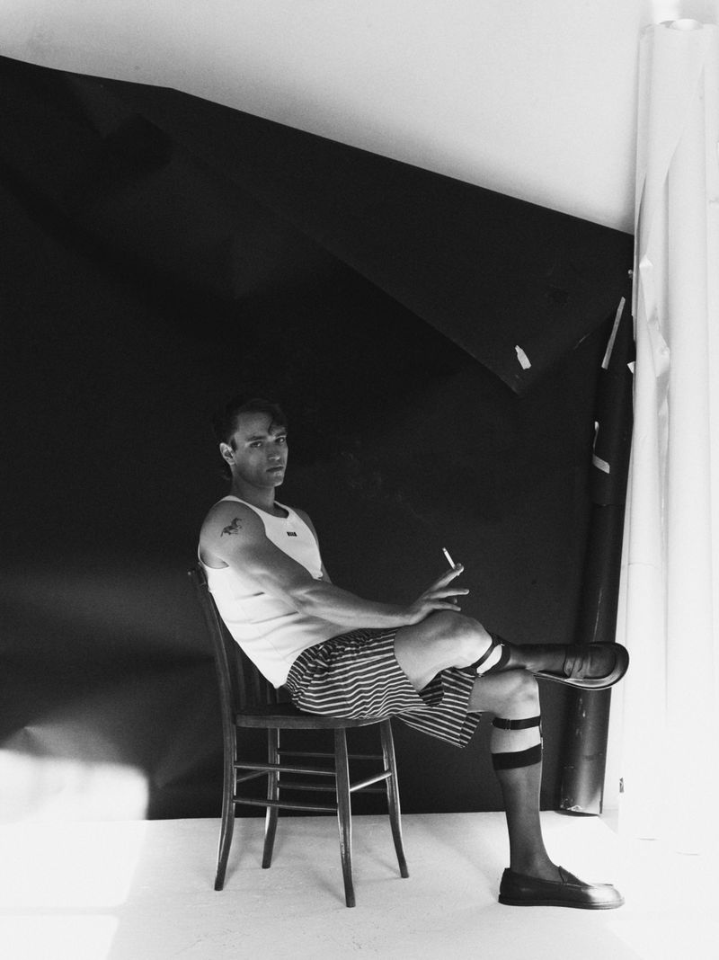 Monochrome portrait of an adult man sitting on a wooden chair against a black backdrop, wearing a tank top and striped shorts, smoking a cigarette.