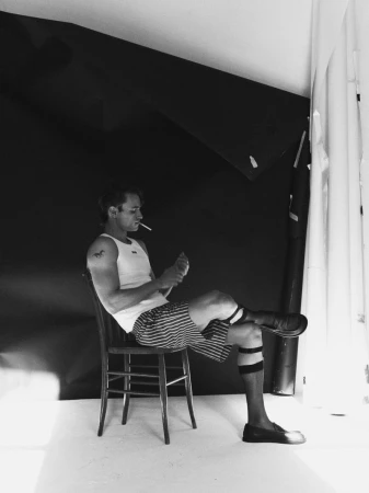 Man sitting on a wooden chair, lighting a cigarette with a lighter, wearing a tank top and striped shorts in a photo studio.
