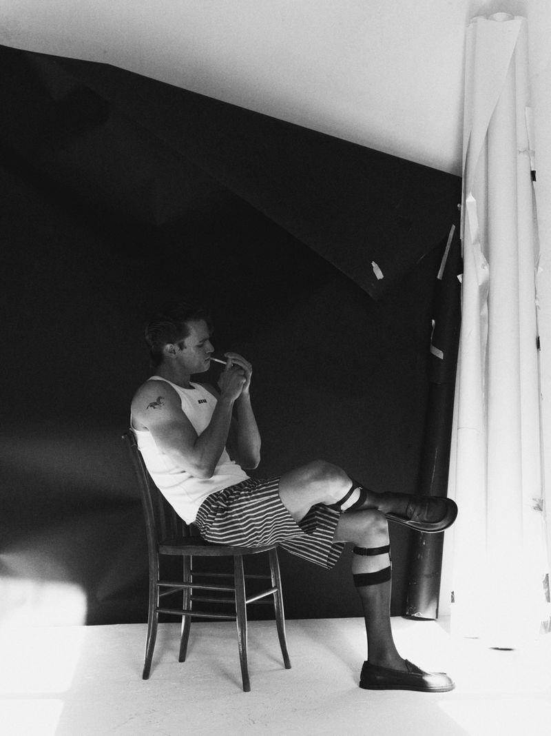 Monochrome studio portrait of a man in a white tank top and striped shorts seated on a wooden chair.
