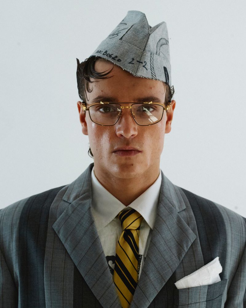 Portrait of a person wearing a grey pinstripe suit, yellow tie, glasses, and a cap, posing against a white background in a studio.