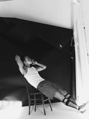Monochrome portrait of a man in a white tank top and striped shorts leaning back on a dark wooden chair.