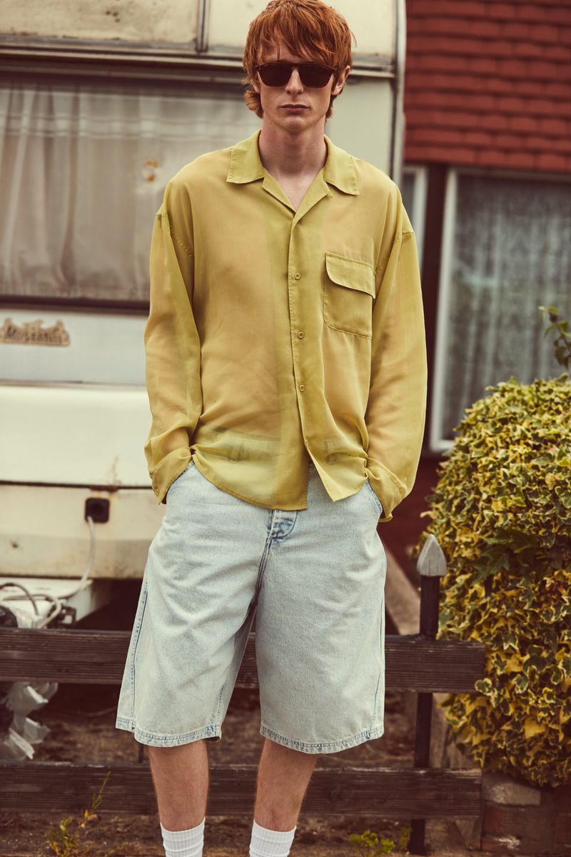 Red-haired man in sunglasses and yellow shirt poses in front of a vintage trailer. He wears jean shorts and white socks.