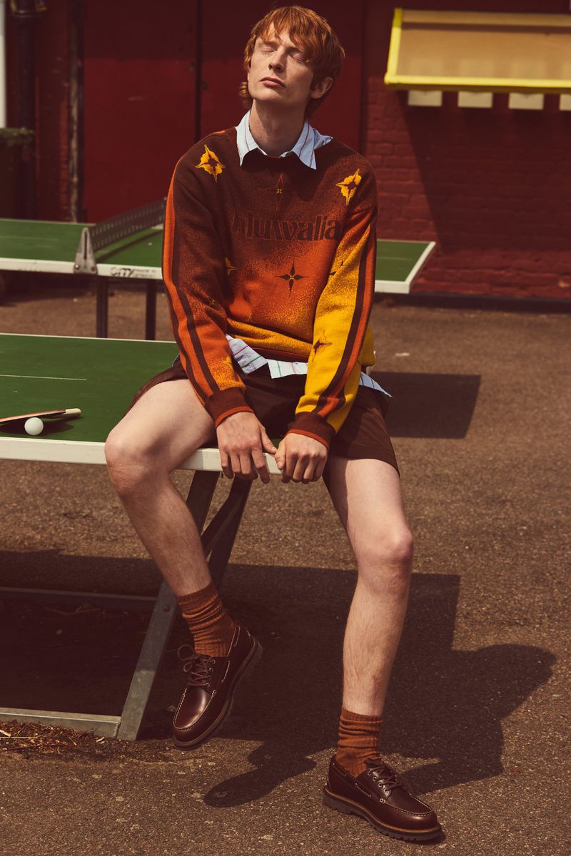 A fair-skinned, red-haired young man sits on a table tennis table outdoors, wearing a patterned sweater, shorts, socks, and dress shoes.