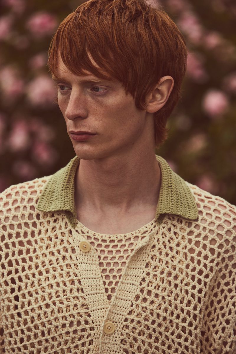 Close-up of a young, red-haired man in a knit shirt with a green collar, standing outdoors amongst flowering plants.