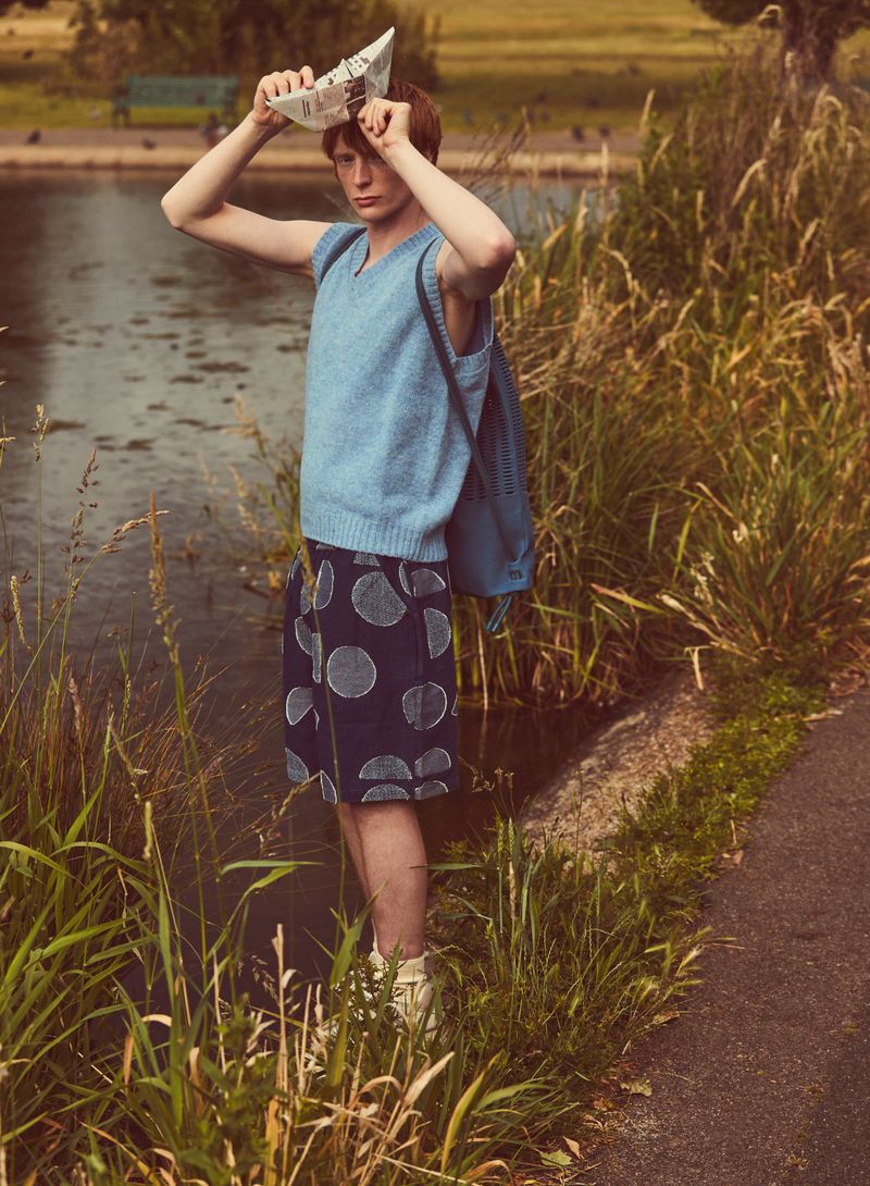 A young adult male in casual clothing stands next to a pond, holding a paper boat over his head in a park setting.