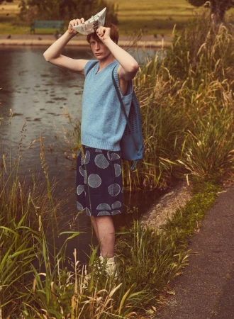 A young adult male in casual clothing stands next to a pond, holding a paper boat over his head in a park setting.