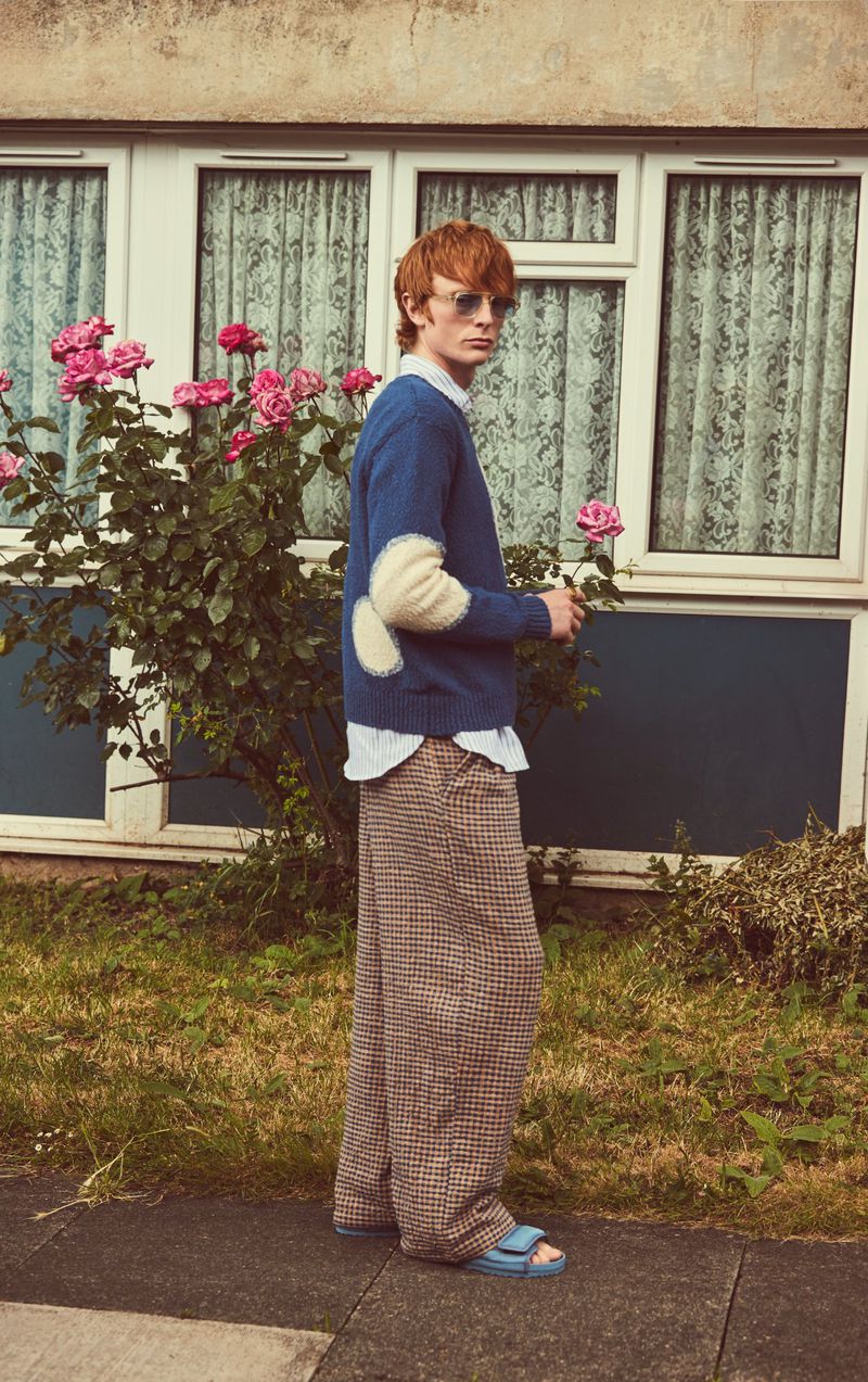 Male model poses outdoors in blue sweater with elbow patches, holding a rose, wearing wide-leg pants, and light blue sandals.