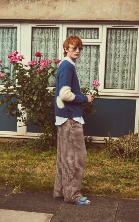 Male model poses outdoors in blue sweater with elbow patches, holding a rose, wearing wide-leg pants, and light blue sandals.