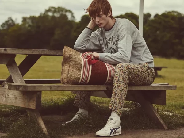 A young man is sitting on a weathered wooden picnic table in a field, wearing a sweatshirt, patterned pants, and hi-top sneakers, looking contemplative.