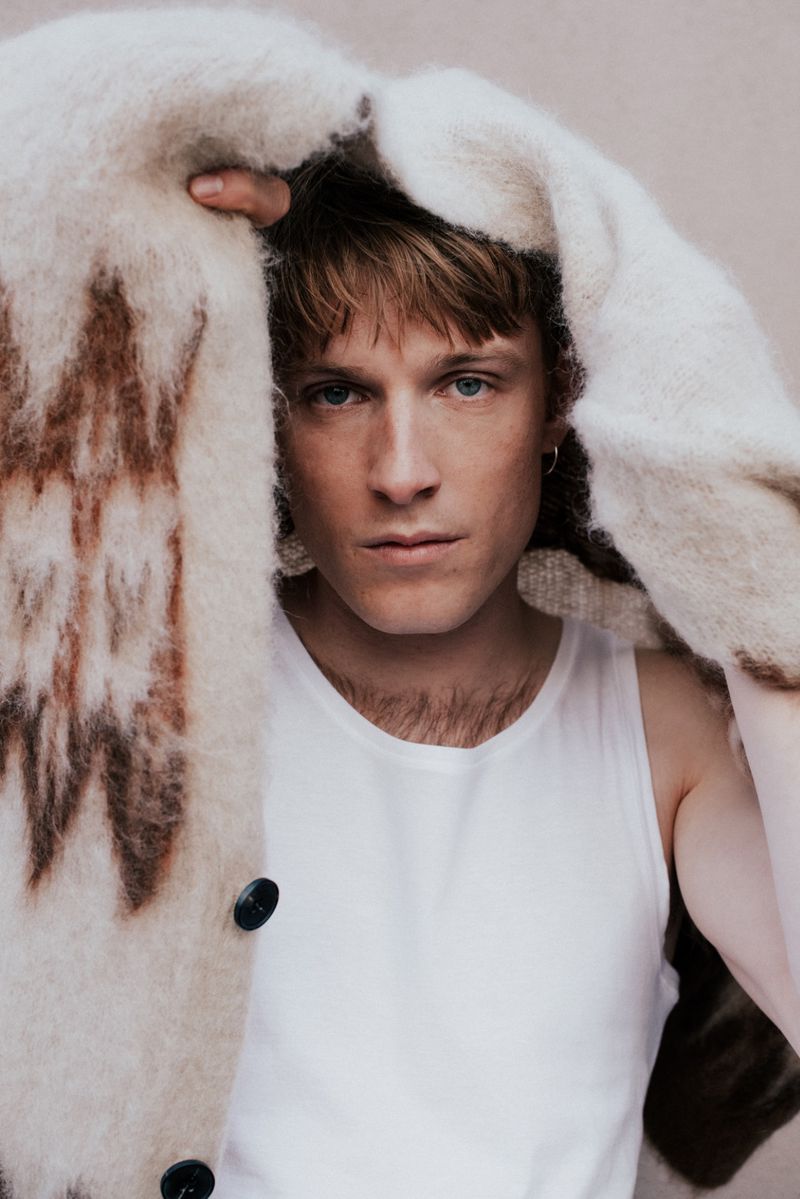 Portrait of a blonde male wearing a white tank top and a patterned beige cardigan, looking directly at the camera indoors.