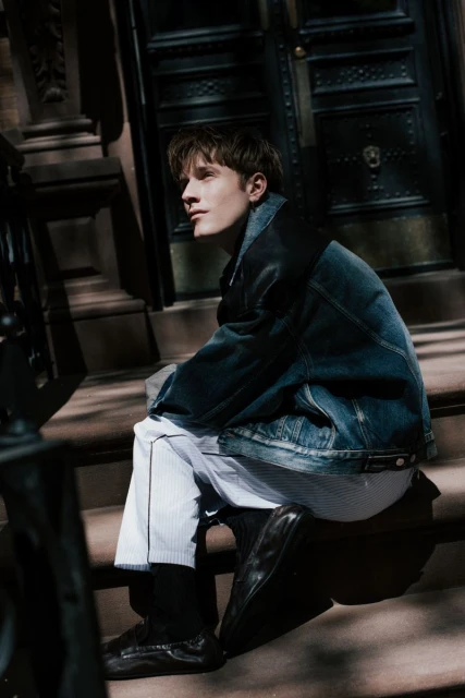 Young man poses on brownstone steps wearing a denim jacket, striped shirt, and black shoes in an urban environment with natural lighting.